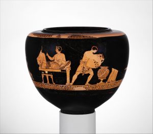 A muscular, enslaved young man pours some liquid into a container on the ground. The presence of reclined men on the couch suggests the room is an andron and a male-only party or symposion is in progress. So, perhaps the storage container is a wine krater, and the enslaved man is pouring water into it, since Greek generally drank diluted wine.