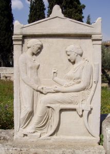 A Funerary tombstone depicts a standing enslaved female attending her enslaver mistress, seated in a finely made chair. The inscription on the top identifies the seated woman's name as Hegeso, implying the social status and wealth of the woman.