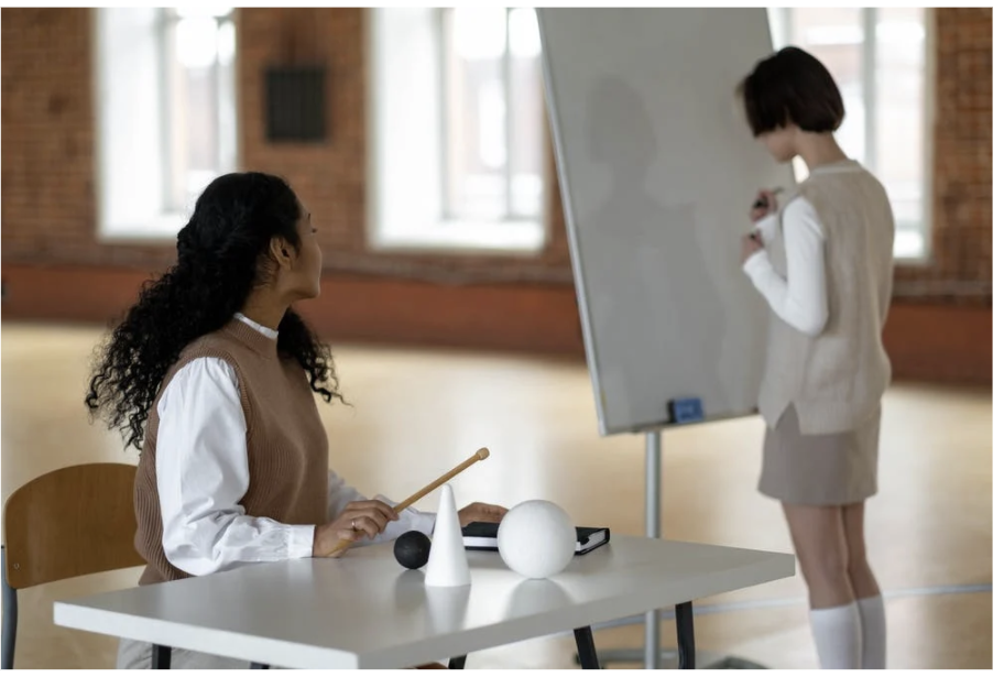 Female teacher is sitting at a desk. Female student is at the front beside the teacher about to write on a whiteboard