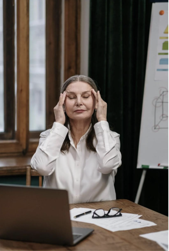 Female teacher sitting at her desk with eyes closed and both hands on her temples.