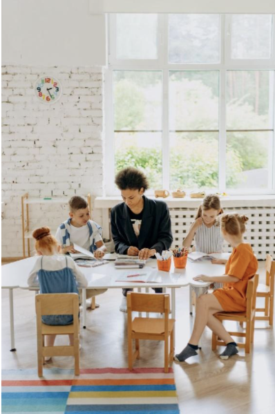 Students sitting on a brown wooden chair looking at their papers. The teacher is sitting at a table with them.