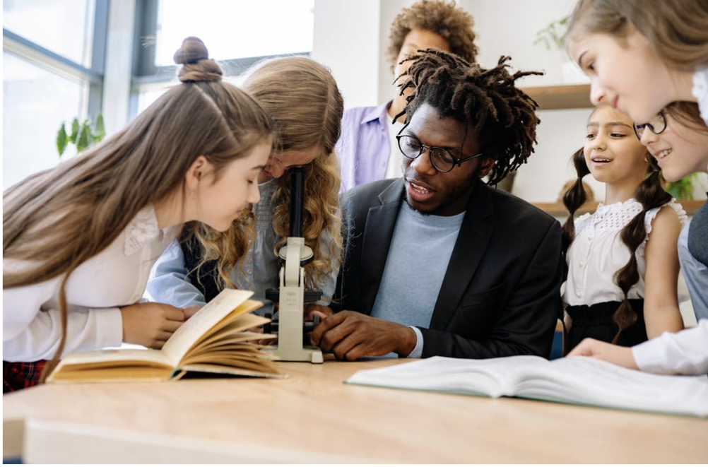 Male teacher sitting at a round table with students surrounding him, one female student looking into a microscope.