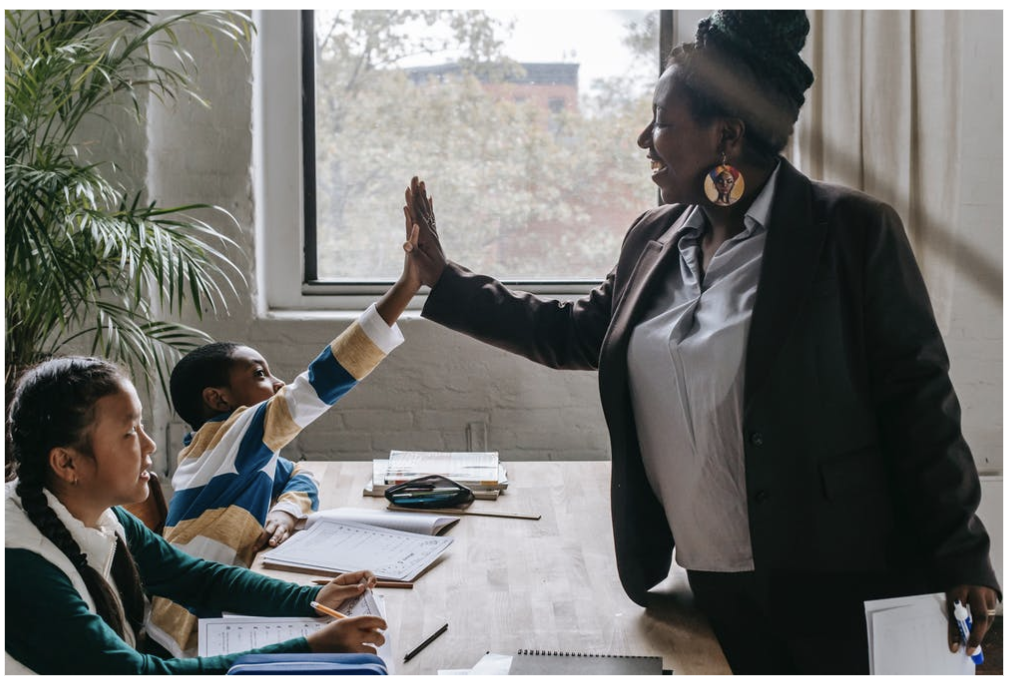 Female teacher high fiving a male student from across the table. Another student is sitting at the table