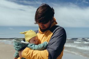 Man in a yellow windbreaker holding a baby in a yellow hat on a cold, windy beach