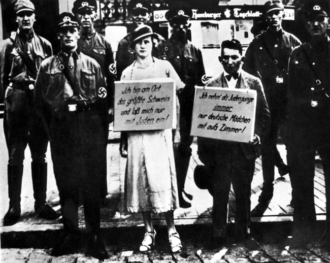 Un homme juif et une femme aryenne sont humiliés publiquement pour leur relation sexuelle en étant forcés de tenir des panneaux en Allemagne, 1933. Les pancartes disent : « En tant que Juif, je n'invite toujours que des Allemandes dans ma chambre! » et « Je suis la plus grosse truie de la ville et je n'ai affaire qu'à des Juifs. »