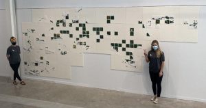 Two people stand in front of a series of beaded tiles mounted on a wall. The beaded tiles form a fragmented map of Lake Nipissing and the French River.