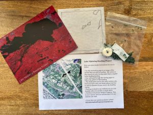 Photograph, taken from above, of contents of a beading kit on a table. The view includes a postcard with a dark lake and red-coloured land with the words 'Lake Nipissing Beading Project' (left), a felt square with water outlined (top), plastic bag of beads with needles and a spool of thread (right) and a paper with a map and instructions (bottom).