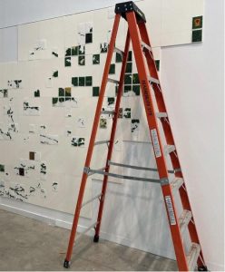 Digital photograph of a ladder standing in front of a white gallery wall with a beaded map in the process of being installed. The map is unfinished and is made up of individually beaded squares that represent different parts of Lake Nipissing.