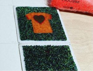 Photograph of two beaded tiles, with green beads tightly stitched together in various patterns depicting water. One tile has an orange shirt and heart beaded into the centre of the water, honouring children who attended and died at residential schools.