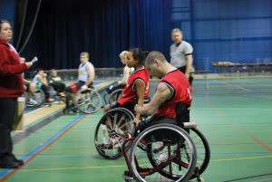 This image is of four wheelchair basketball athletes in BB chairs, wheeling to the sidelines; one woman, perhaps the coach, awaits the two athletes in the red tank top; the other two athletes have blurred spectators in their background; the referee wears grey; the gymnasium floor is bright green.