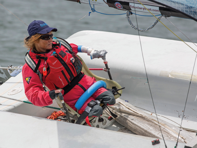 This image is of a quad amputee woman in white sailboat, working with the sail, leaning to the right; red life jacket, blue baseball cap, long hair blowing in the wind; water in the background.