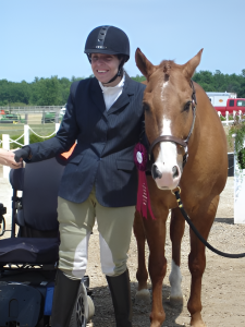 This image is of a female para-athlete in equestrian riding uniform of black hat, blue blazer, beige pants, and boots; she is smiling and standing between her dark blue chair and a chestnut-coloured horse with a white nose; field and trees in the background.
