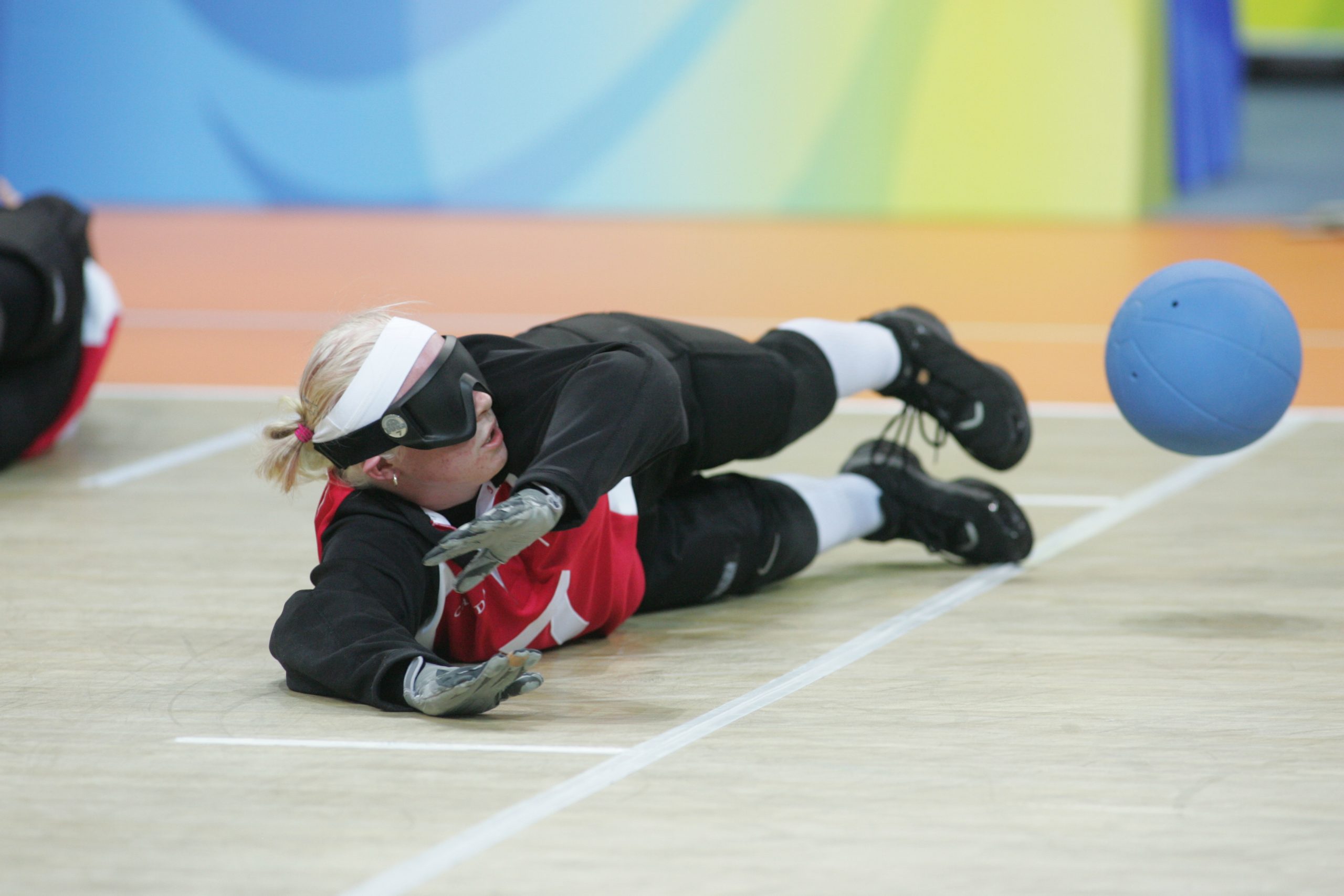 This image is of a goal ball female athlete in a side lying defensive position preparing to stop the large blue goal ball coming towards her; blond hair, white headband, black tights and long sleeved t shirt, red tank top over the black t shirt, white socks, black athletic shoes; grey gymnasium floor, while lines on the floor, mats in the background.