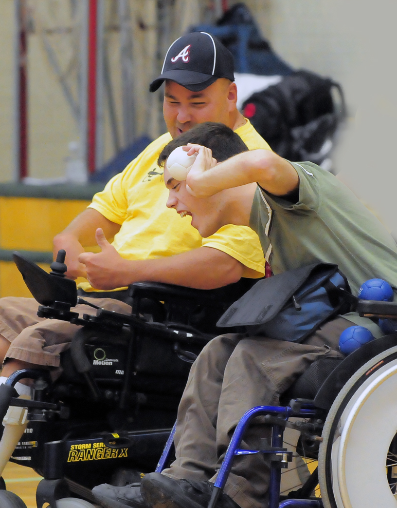 This image is of two male wheelchair users, side by side; the smaller male, dark hair, in a green t-shirt, holds a small white ball, preparing to throw; the larger male in a yellow t shirt and a blue baseball cap is next to the smaller male preparing to throw; blurry gymnasium wall in the background.