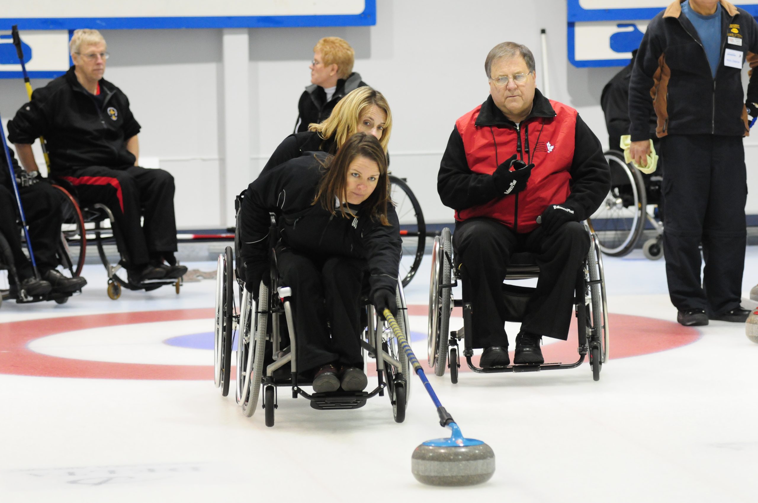 This image is of six wheelchair curling athletes, all using wheelchairs; one other person in the upright position; all on a curling rink surface, white with a blue circle inside a larger red circle; two women and one man in the foreground; the most foregrounded woman is pushing the curling rock with the arm extender.