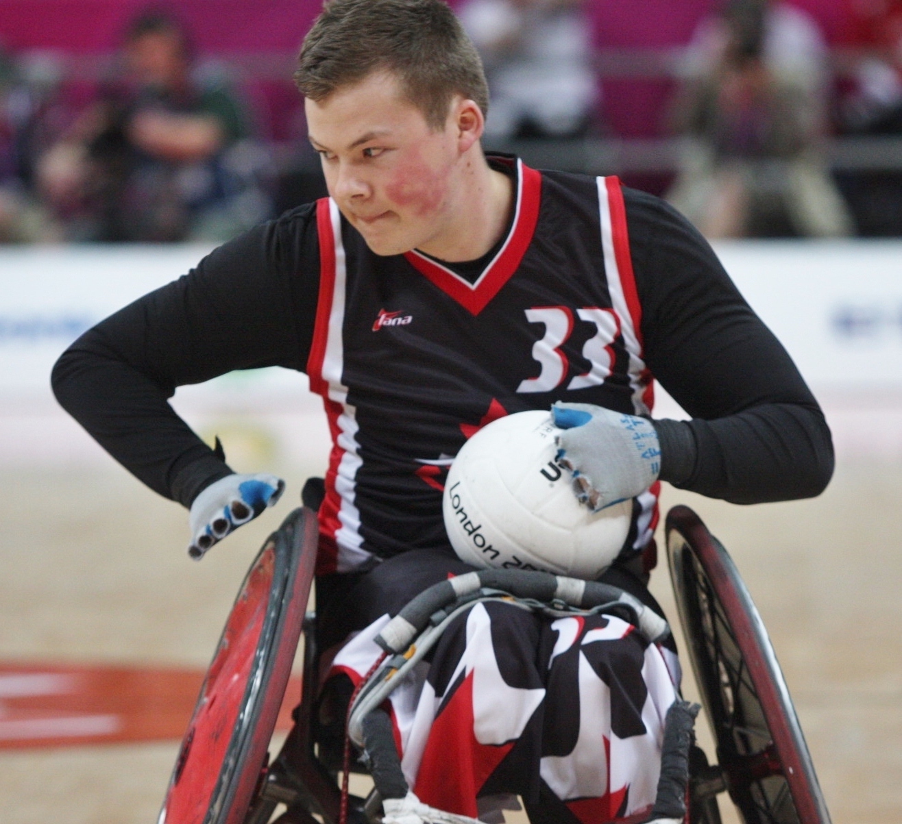 This image is of a male wheelchair athlete; chair has court wheels with red inserts; athlete has short light brown hair, wears black with a pinnie with number 23 in white; he is looking to the side, carrying a round, white ball on his lap; he wears grey fingerless gloves; blurry audience in the background.