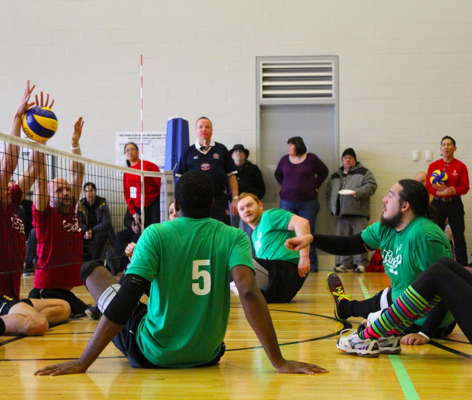 This image is of a sitting volleyball game in a gymnasium with spectators viewing while standing in front of the far wall; there are five athletes in green on the right side of the image looking over the net; there is a blue and yellow ball suspended over the net; the three athletes wearing read on the opposite side of the net are blocking the ball; an official wearing black and using a whistle stands at the blue post near the far wall.