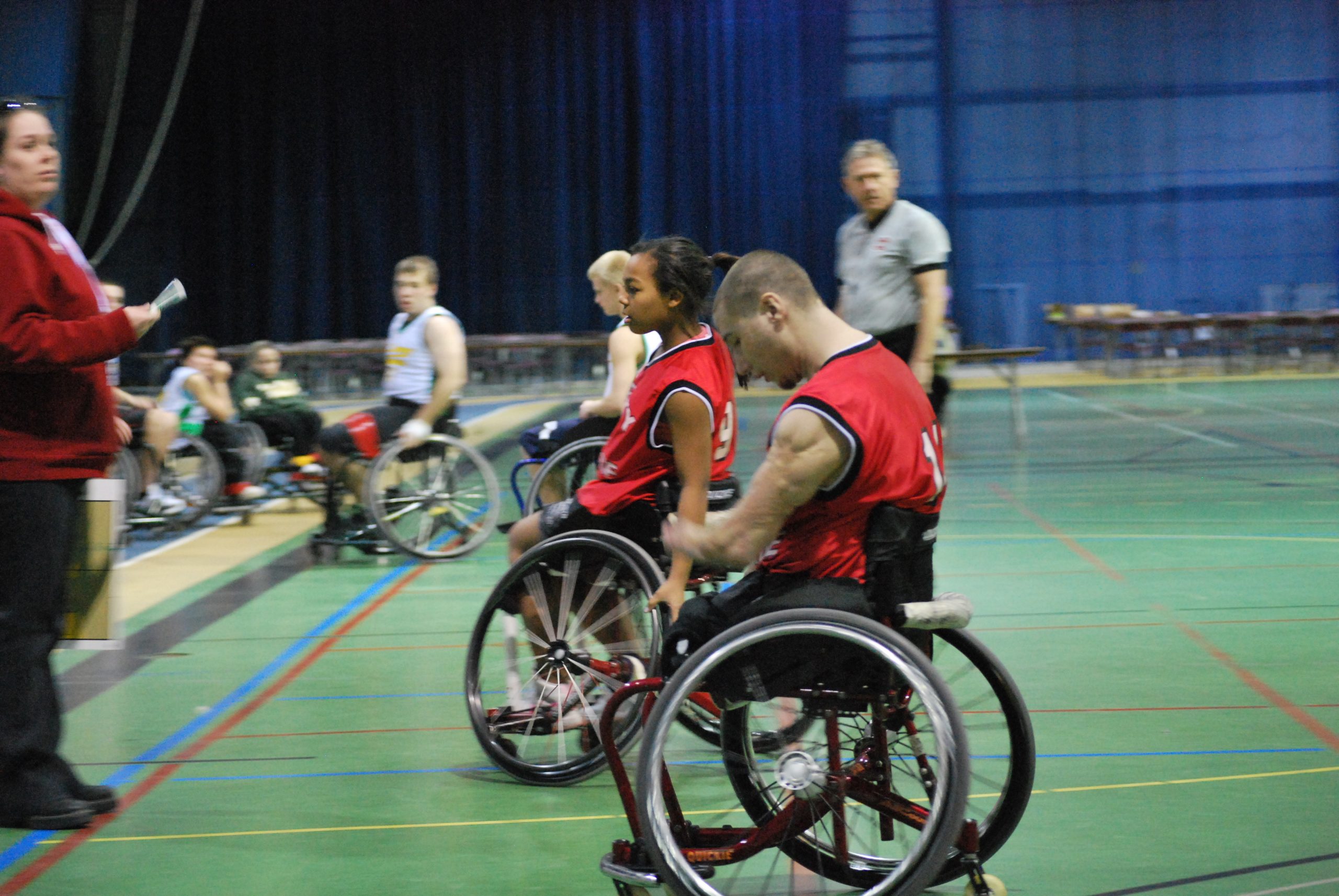 This image is of four wheelchair basketball athletes in BB chairs, wheeling to the sidelines; one woman, perhaps the coach, awaits the two athletes in the red tank top; the other two athletes have blurred spectators in their background; the referee wears grey; the gymnasium floor is bright green.