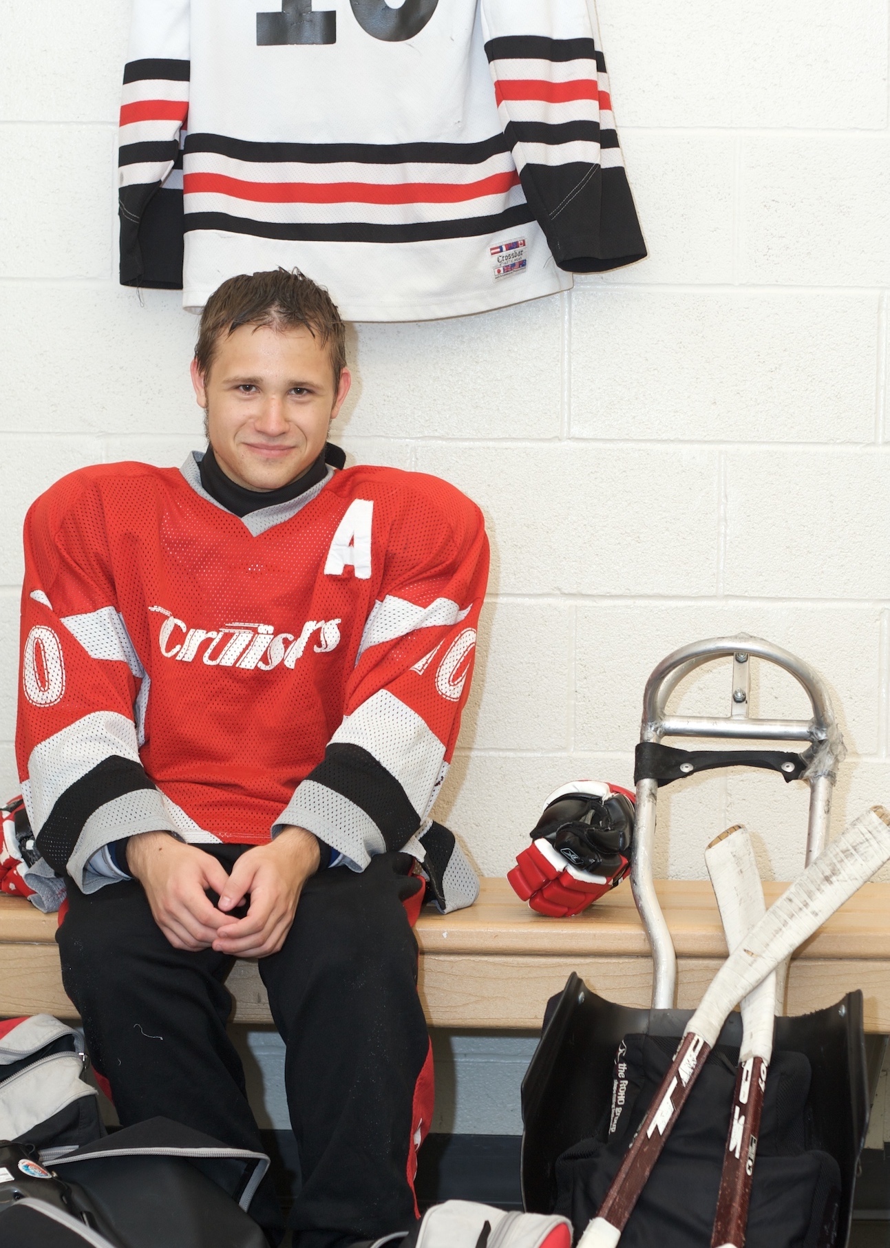 This image is of a young male para-ice hockey athlete sits in the dressing room against the wall, on a bench, next to his sled; his gloves are on the bench and his sticks are crossed over in front of his sled; his brown hair looks damp; he wears a red hockey jersey with the team name in white letters and a large A on the upper shoulder of the jersey; there is a white hockey jersey hanging on the wall behind him.