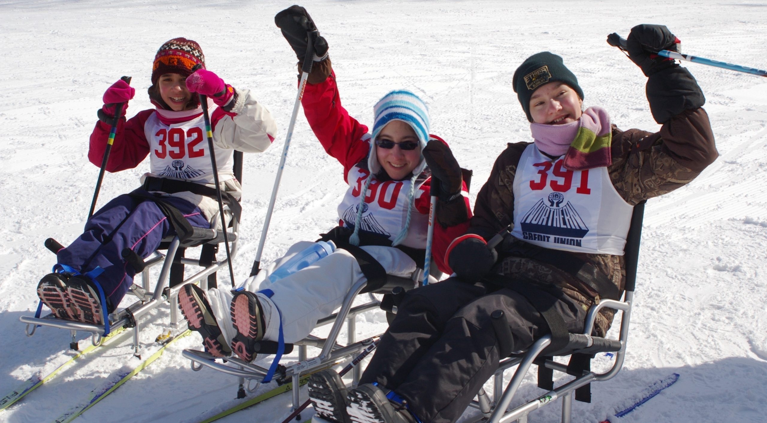 This image is of three people in sit skis on snow; all three wear winter clothing and hats and have pinnies with numbers on them; all three are using ski poles; the person in the foreground has one pole lifted; the person in the middle has one pole lifted; the third person has both poles in the snow.
