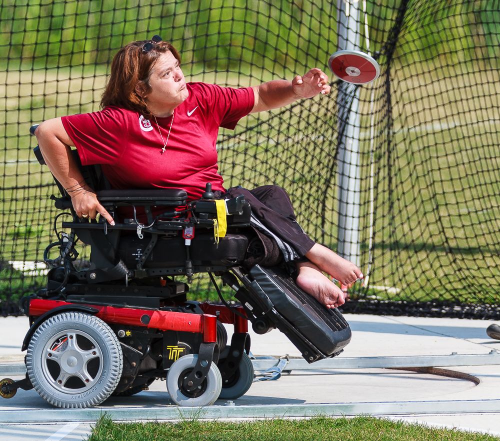 This image is of a female wheelchair athlete throwing a discus; her arm is outstretched as she releases the discus; she wears a red t-shirt, has neck length brown hair and is wearing a head band; she uses one arm to brace herself against her chair; she has bare feet; there is black discuss netting and blurry greenery is in the background.
