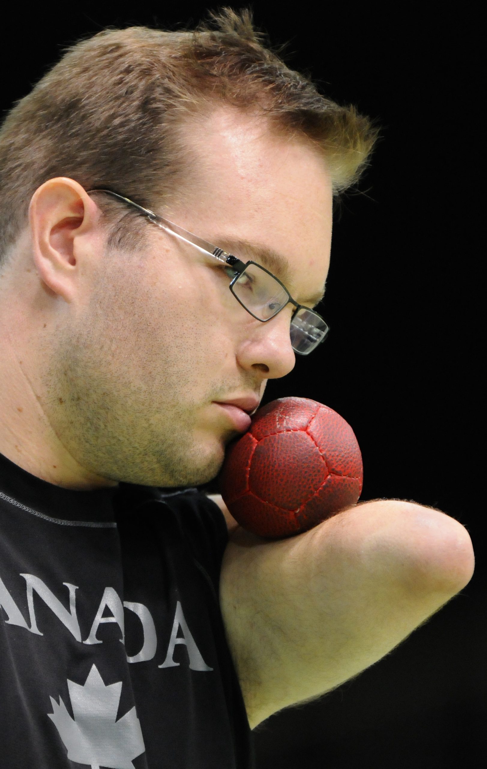 This image is of a male upper arm amputee holds a red ball between his stump and his cheek; he has short brown hair, glasses, and some beard stubble on his face; he wears a black t shirt with the word CANADA in white letters.