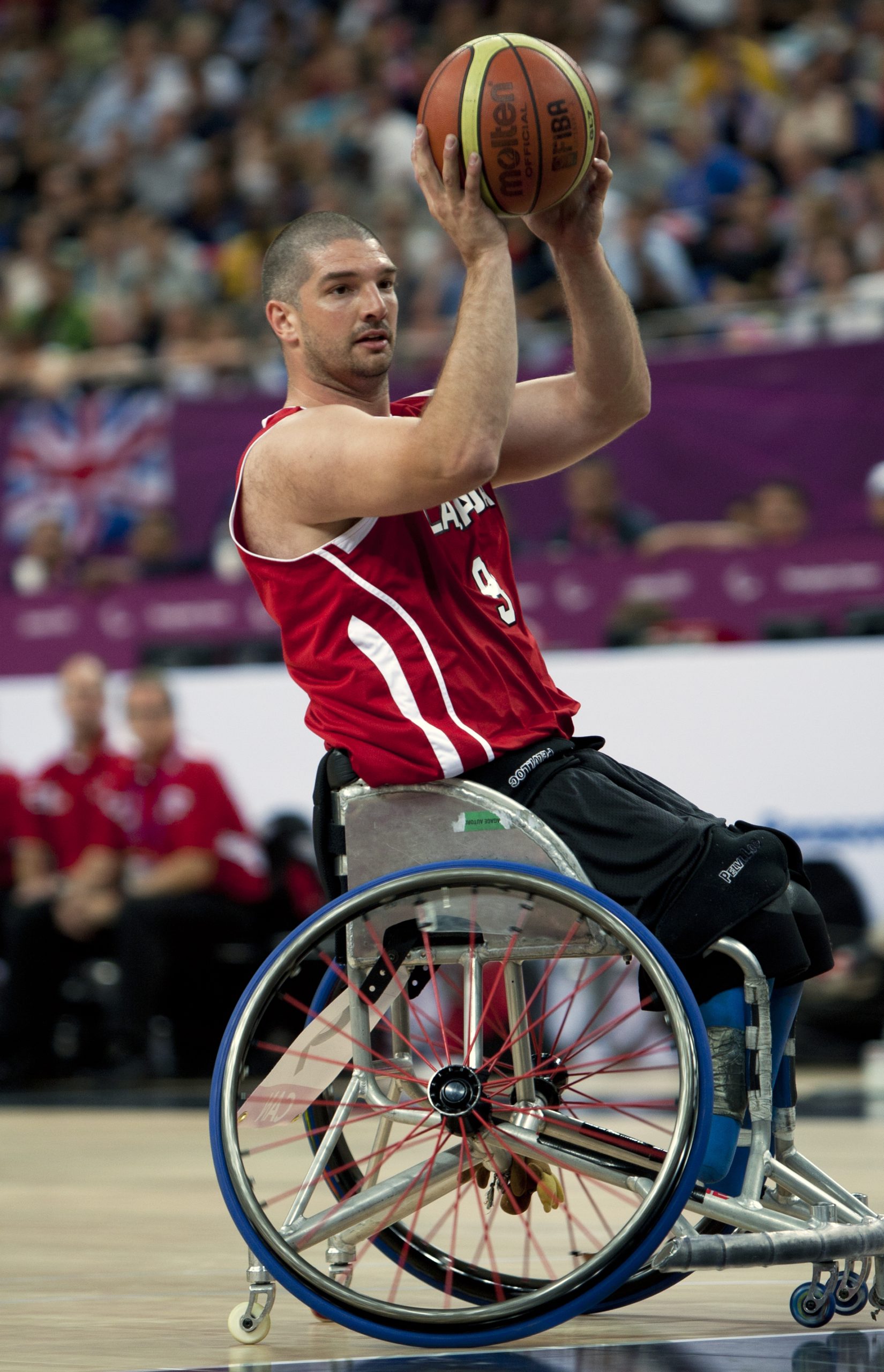 This image is of a wheelchair basketball male athlete sitting high in the wheelchair, with a basketball poised to shoot; the athlete’s hair is short and dark; he wears a red tank top and black pants; the background is a blurry spectator audience.