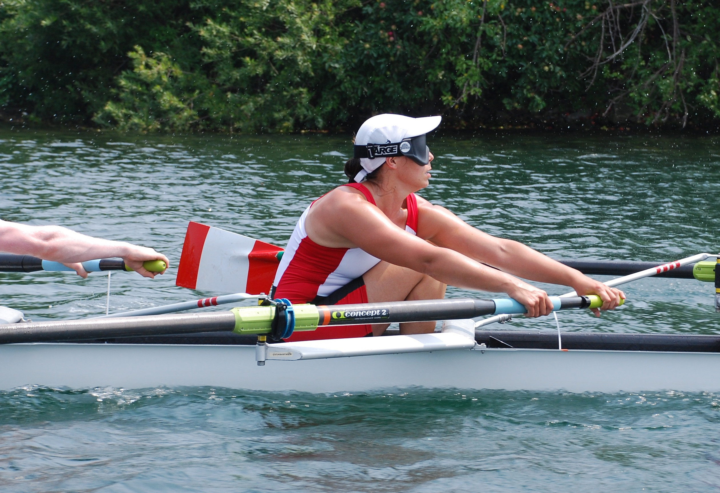 This image is of an athlete in a rowing shell; the athlete hods two oars and is in the process of pulling on the oars; the boat is on the water with greenery in the background; the athlete wears dark glasses, a white baseball hat and a red and white rowing singlet; the athlete’s arms are outstretched, and knees are bent.