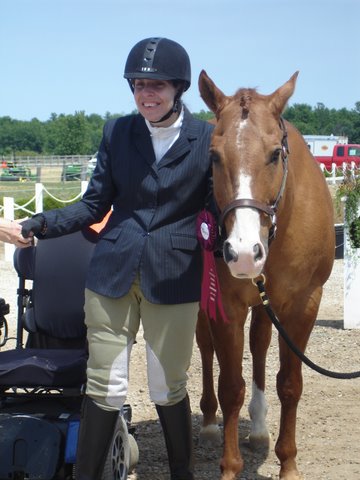 This image is of a female para-athlete in equestrian riding uniform of black hat, blue blazer, beige pants, and boots; she is smiling and standing between her dark blue chair and a chestnut-coloured horse with a white nose; field and trees in the background.
