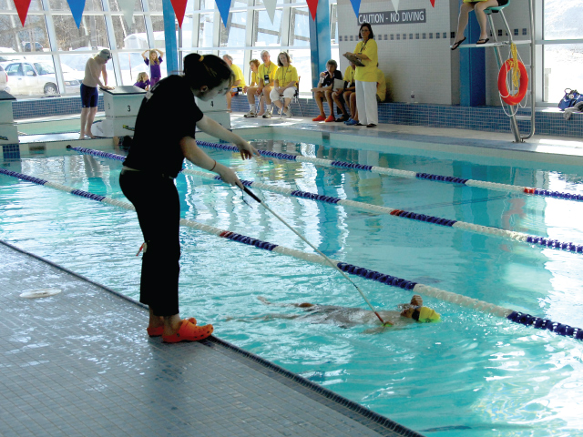 This image is of a para-swimmer in a swimming lane in the water near the side of the pool with a coach-observer on deck beside the swimming lane using a tapping wand; there are several people wearing yellow in the background; the pool has glass windows as walls, the lane markers are blue and white, there is a red lifesaver ring hanging off a lifeguard chair, the tapper wears black clothes and red crocs, the water appears to be blue.