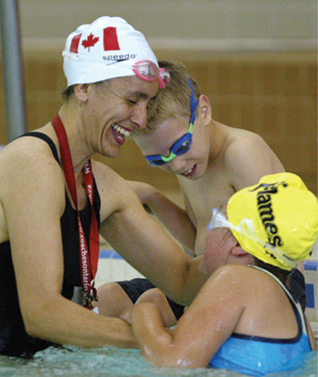 This image is of an adult female swim instructor with white swim hat with a red maple leaf on it is smiling and interacting with two children in a swimming pool; the girl wears a blue swim suit, goggles, and a yellow swim hat with the word ‘flames’ in black letters; the boy wears black trunks and blue and green goggles; all three people are close together in the water; there is a blurry wall back ground and one side of a swim ladder near the adult female.
