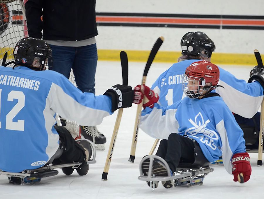 This image is of three para-ice hockey athletes are on the ice in sledges; each wears blue hockey jerseys and hold sledge sticks; the two larger athletes wear black helmets and the smaller athlete wears a red helmet; there is a partial image of a person in an upright position to the side; the ice is white and there is a background of white hockey boards with a red and black stripe.