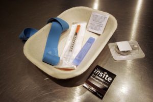 Photo of a safe injection kit from Insite Supervised Injection Site in Vancouver, BC. The kit includes a thick medical rubber band, sterile needle syringe, a rubbing alcohol prep pad, and a pipette. These items are in a disposable cardboard plate. Outside the plate is a small metal spoon for melting drugs and a matchbook with "insite" and phone number on it.