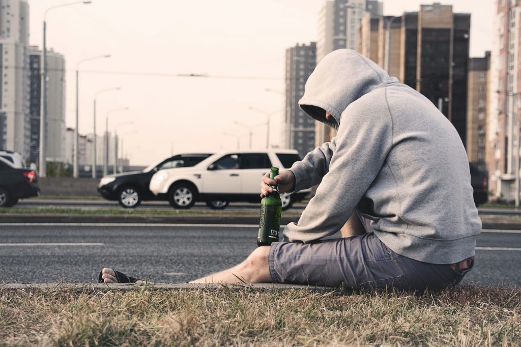Person in gray hoodie and shorts sitting on the sidewalk on the side of a busy road, holding a bottle of beer in their had in Moscow, Russia
