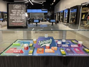Display cases and touch screens in a One Plant cannabis dispensary. On the glass case is a sign that says, "Enter only if you are 19 years or older."The displays show different forms of cannabis, including flower, edibles, vape, and preroll.