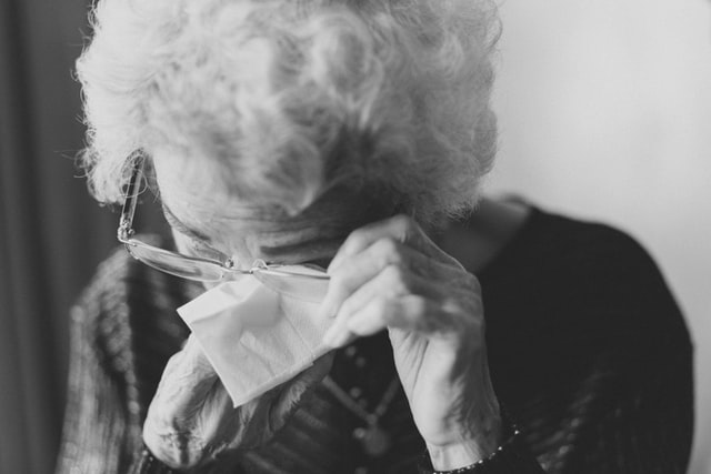 Elderly woman with glasses crying and wiping eyes with tissue 