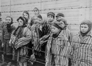 group of children standing behind a barbed wire fence in Auschwitz wearing striped adult-sized prisoner jackets