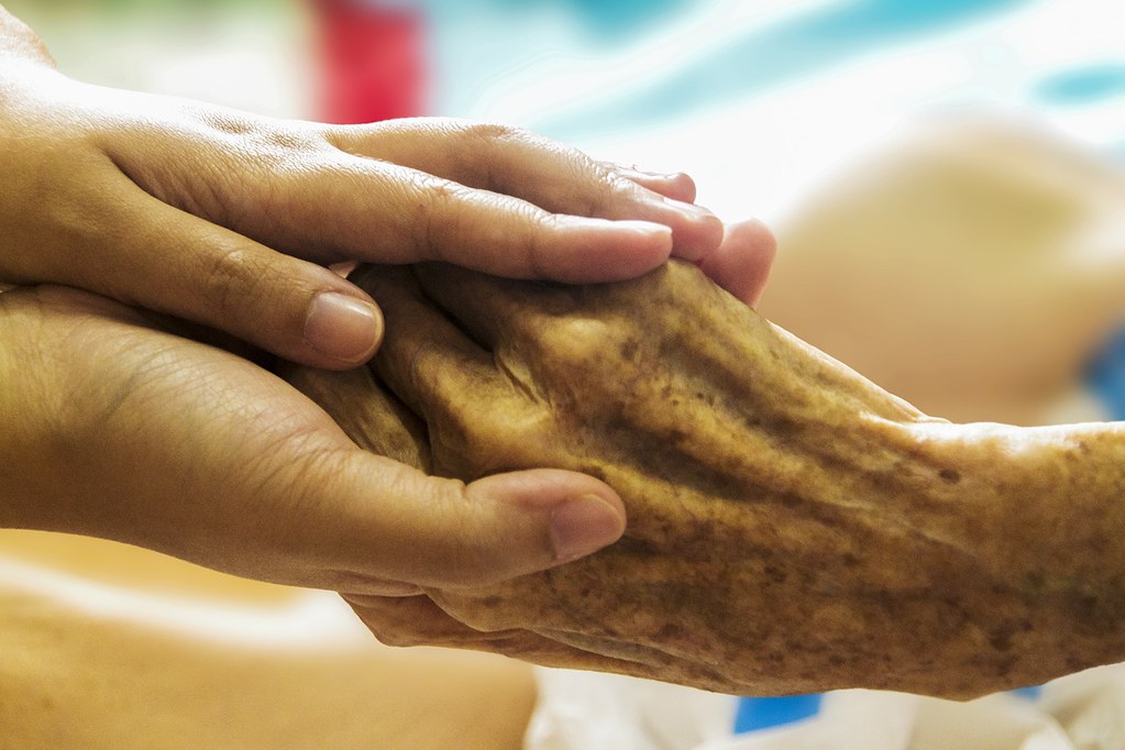 palliative care worker's hands holding the age-spotted hand of a patient