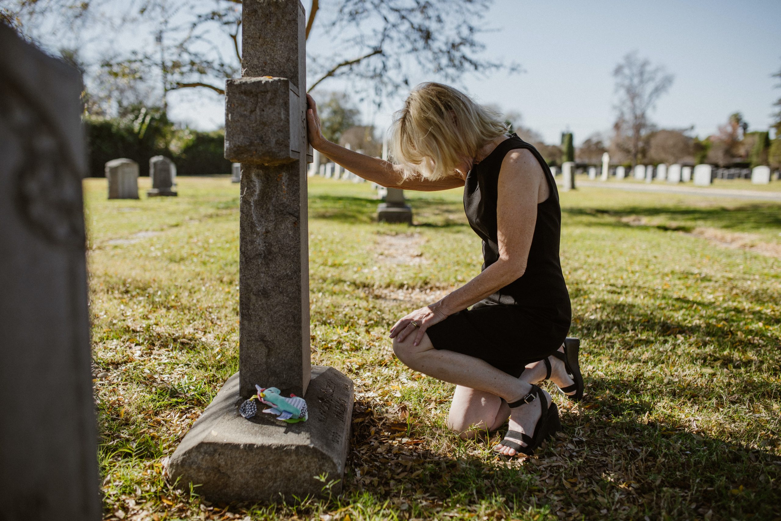 woman in dress kneeling on grass with arm on a gravestone. The gravestone is in the shape of a cross, and there are flowers at the base of it.