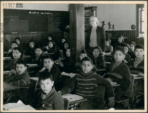 a classroom of Indigenous students in a classroom at a residential school. A white female teacher stands amongst seated students. All students have short hair.