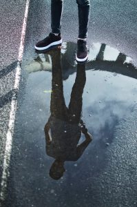 Photo of a man standing on the road in front of a puddle of water that shows his reflection.