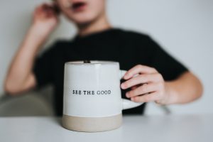 Photo of a person in a black shirt holding a white mug that reads, "see the good."