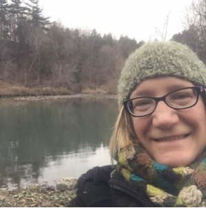 Jennifer Woodill, a white woman with short blond hair wearing a green beanie hat and scarf in front of a lake and trees along the lake.
