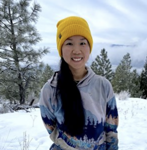 Teresa Lee, an Asian woman with long black hair wearing a yellow beanie hat in front of trees along the snow-covered trail.