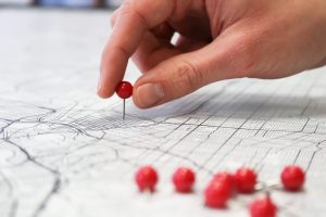 a white person's hand holds a red tack between their fingers as they push the pin into a white paper map. In the foreground there are other red pins already pushed into the map.