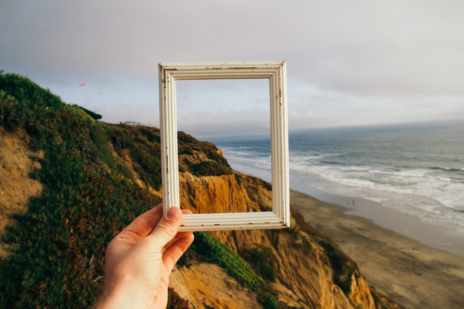 Photo of a hand holding up a frame in front of a backdrop of cliffs and the ocean.