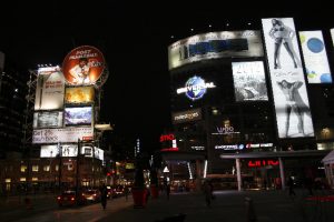 Night scene of a busy intersection with sides of skyscrapers all showing different sizes of advertising from a few feet tall to multiple stories.