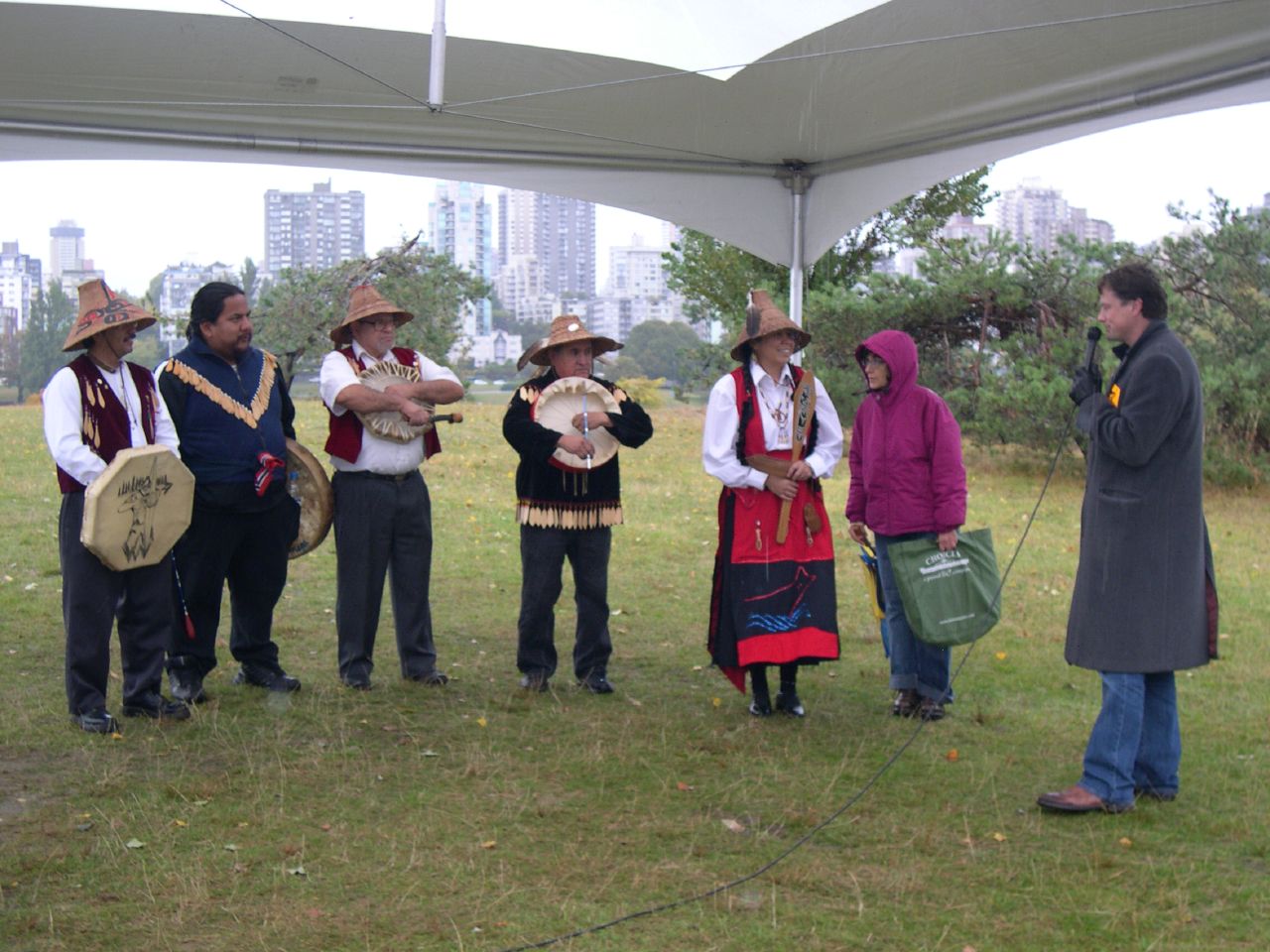 This photo shows a number of people under a tent on a grassy lawn, with bushes to the right. In the background are high-rise buildings. One man is holding a mic. Five people are dressed in traditional clothing and four of them are holding hand drums. Four of the performers are wearing bell-shaped hats woven from cedar bark.