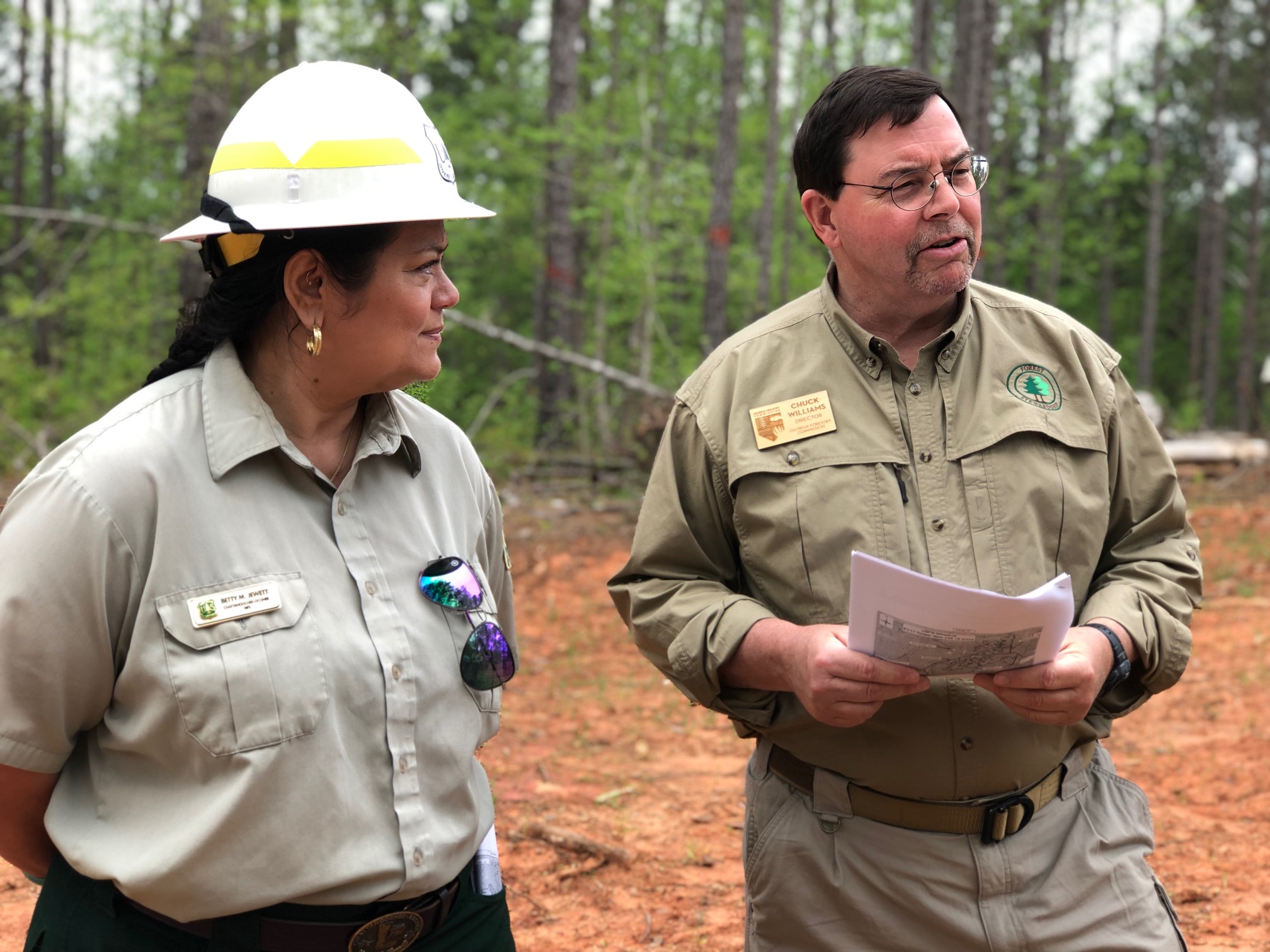 Two people in foresters' uniform standing in front of a forest. One is looking at people outside the photograph and speaking. He is holding papers in his hand. The other, a woman, is listening to him. The mood is friendly and respectful.