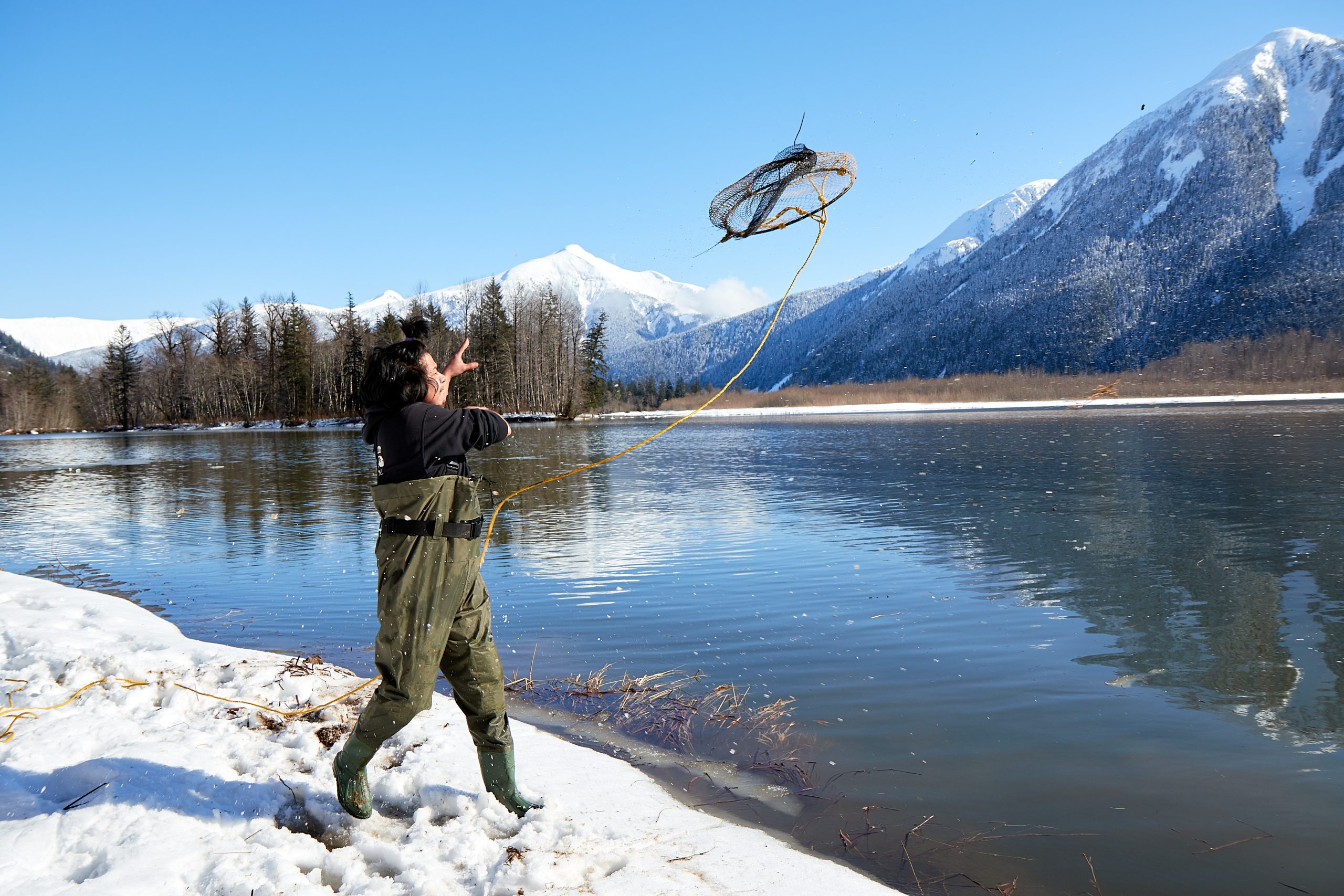 A person wearing chest-high waterproof pants and rubber boots hurls a conical net on a rope into the water. Snow-capped mountains frame the opposite shore.